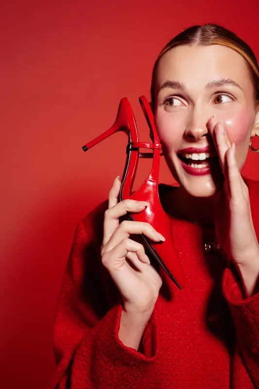 Happy woman in red sweater holds red high heel shoe near face, smiling and whispering. Bright red background creates vibrant mood. Concept for fashion, style, and joyful emotions.