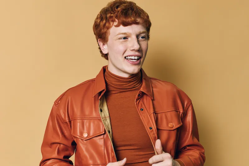 Smiling young man with red hair wearing brown leather jacket and orange turtleneck standing against beige background. Portrait of happy confident male with joyful expression and casual style.
