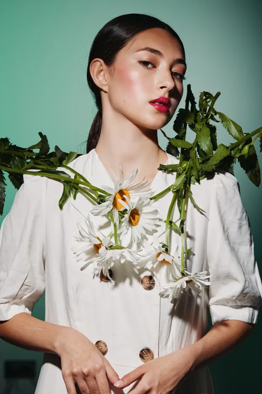 Young woman with bright red lips and fresh flowers around her neck in a minimalist outfit. Fashion and beauty creativity concept with natural elements on soft green background.