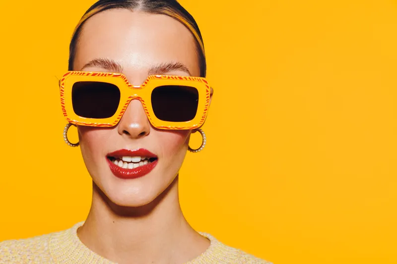 Young woman with bright orange sunglasses and red lipstick poses against yellow background. Fashion portrait with trendy accessories and vibrant colors for summer style concept.