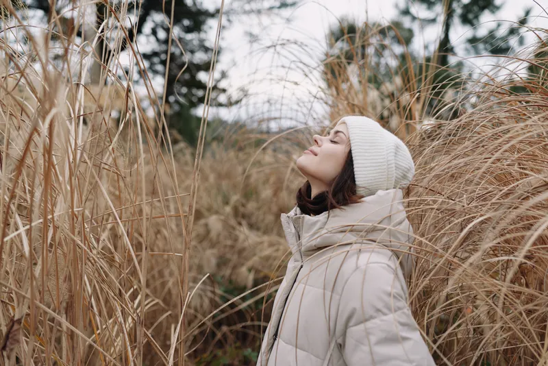 Woman outdoor field grass winter coat beanie stands among tall grasses eyes closed savoring chilly air and peaceful nature in a quiet autumn landscape soft light and calm mood