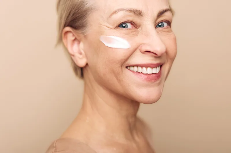 Smiling mature woman applies face cream on cheek with soft beige background. Concept of skincare, beauty, antiaging, wellness, natural glow, and selfcare for healthy skin and confidence.