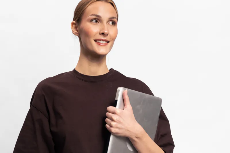 Smiling young woman holds laptop close to chest, looking away with hopeful expression. Casual outfit and clean white background, concept for technology, education, and success.