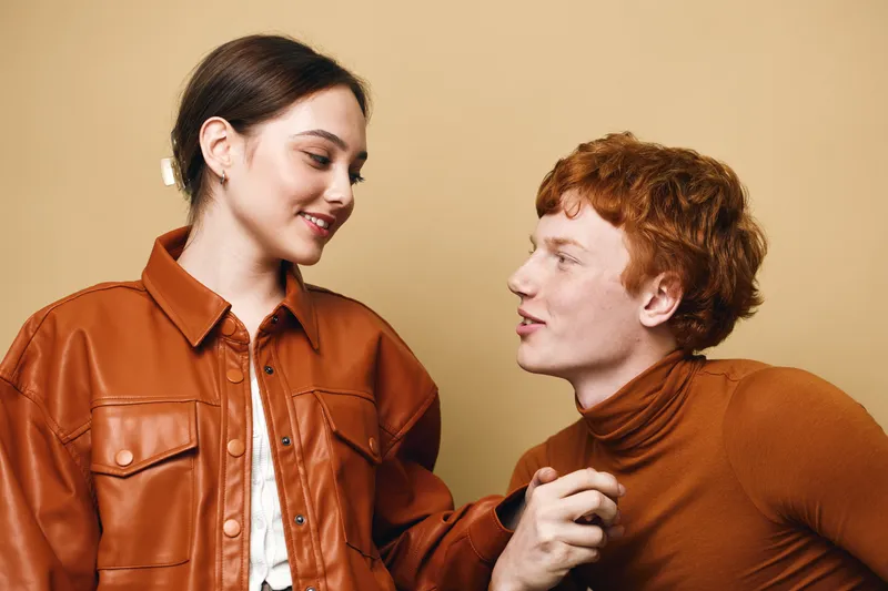 Young woman and man with red hair wearing brown and orange outfits looking at each other with friendly expressions and warm interaction on beige background.