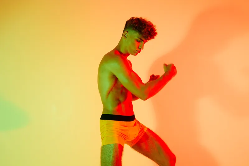 Young muscular man posing shirtless in colorful lighting with flexed arms showing strength and fitness in studio. Male athlete with curly hair demonstrating body power and health.