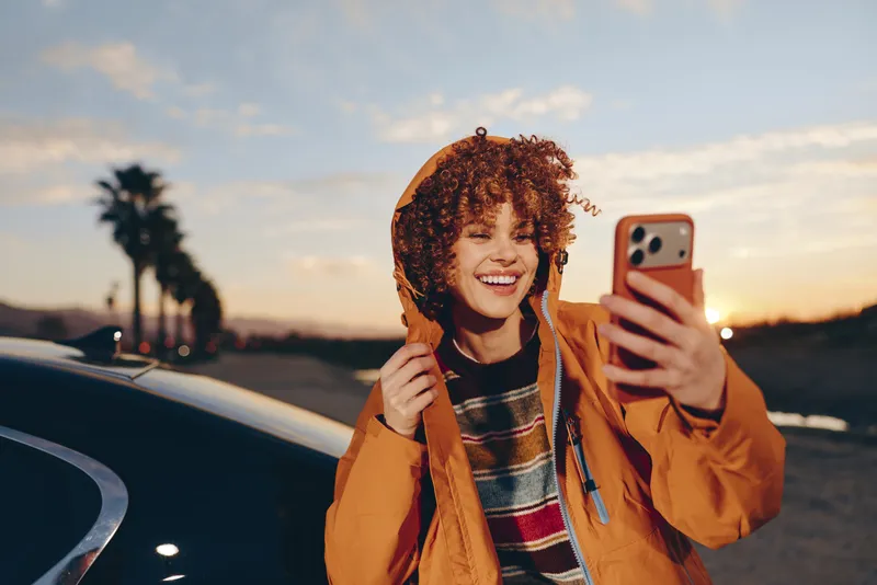Woman selfie smartphone sunset portrait outdoors lifestyle curly hair coat, smiling and holding a phone near a car by the roadside at dusk, warm orange jacket creates a casual travel vibe