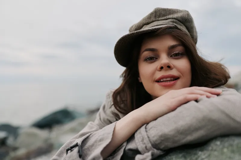 Young woman with brown hair and neutral makeup wears vintage cap and cozy jacket outdoors. She leans on a rock with soft smile, cloudy sky background. Concept of casual autumn style and nature.
