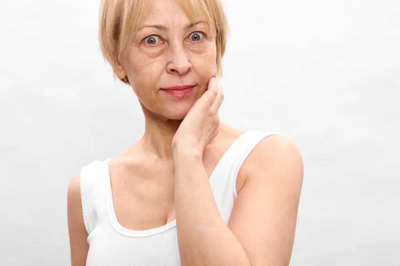 Elderly woman portrait, hand touching face, gentle calm expression, looking at camera, white background, short blonde hair, studio light, soft skin texture, close up, subtle smile