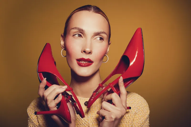 Young woman with red lipstick holds red high heel shoes near face, wearing beige sweater and hoop earrings. Studio portrait on warm brown background with fashion and style concept.