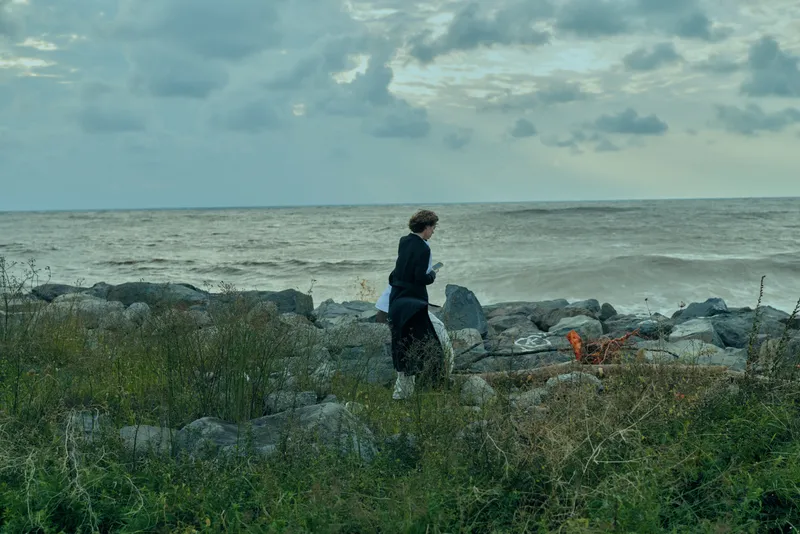 Woman on the windy coast walks among rocks and driftwood, sea in distance, cloudy sky, outdoor solitude and rugged landscape by the shore.