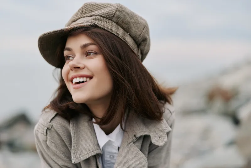 Smiling young woman wearing beige hat and coat looks away outdoors. Natural light highlights her joyful expression and casual style. Concept of happiness and lifestyle.
