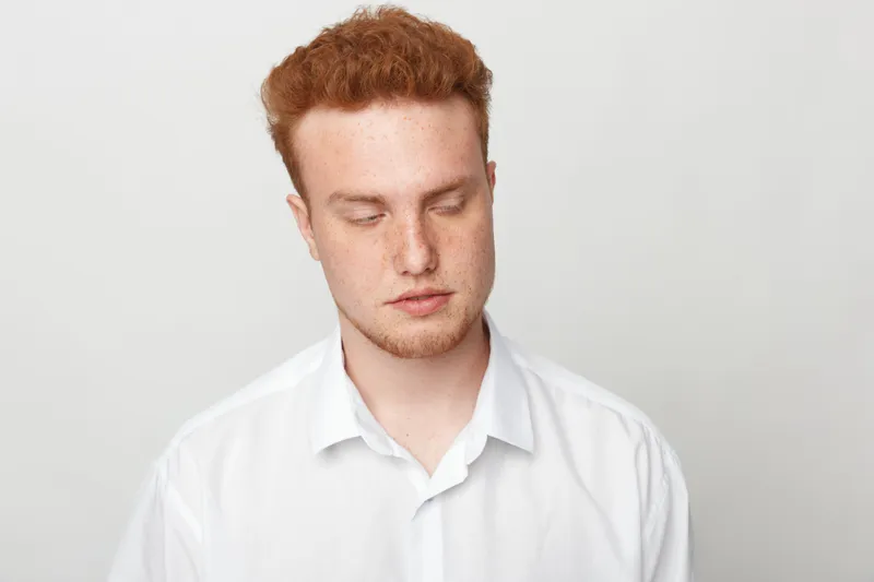 Young man with red hair and freckles wearing white shirt looks down with closed eyes on plain light background. Concept of contemplation, calmness, and introspection in studio portrait.