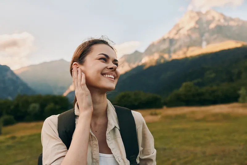 Young woman with brunette hair smiles happily while hiking in a mountain valley during golden hour, wearing a beige shirt and carrying a black backpack.