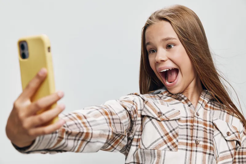 Teenage girl with long hair taking a selfie with smartphone, smiling and happy, wearing plaid shirt against plain background for social media and communication.