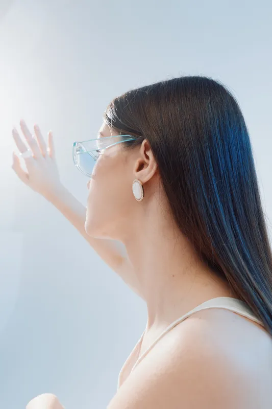 Young woman with long hair wearing transparent glasses and earrings looks out a bright window. Soft natural light highlights her beauty and fashion style in a minimalistic setting.