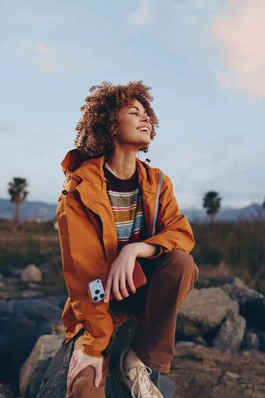 Woman lifestyle outdoors, smiling woman with curly hair and orange jacket, crouches on rocks, holding smartphone, looking upward at mountains at sunset, travel moment in nature