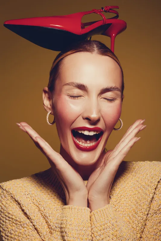 Happy woman with closed eyes and red lipstick laughs joyfully while balancing a red high heel shoe on her head. Concept of fun, fashion, and playful mood in studio portrait.