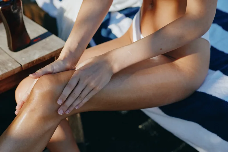 Woman relaxes outdoors by water, sitting on a striped towel, hands resting on legs, wearing swimsuit, warm sunlight highlights skin, candid poolside mood and leisure lifestyle in a calm, carefree