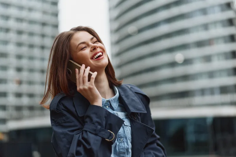 Happy woman with long hair talking on smartphone outdoors in city, wearing blue coat and smiling with eyes closed, urban lifestyle and communication concept.