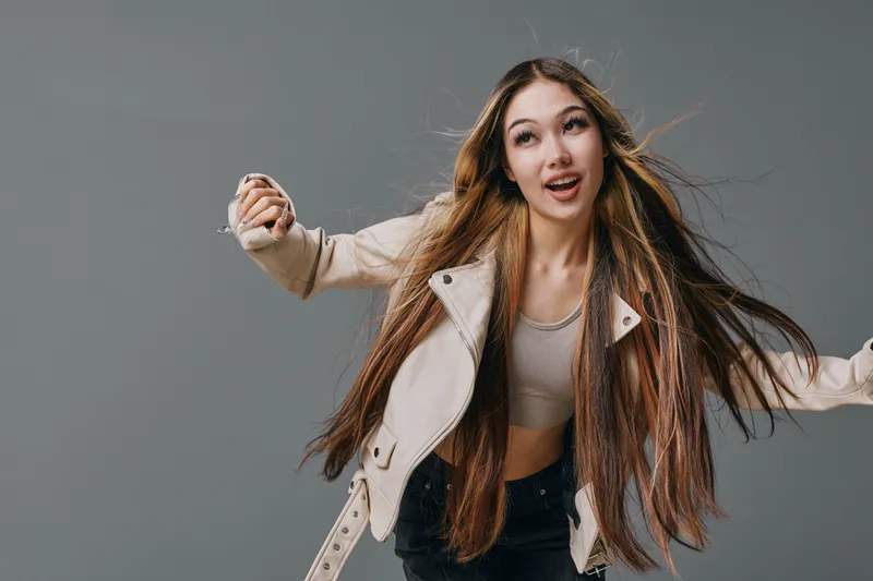 Young woman with long hair wearing cropped top and beige jacket posing with dynamic movement on gray background in studio for fashion and portrait photography.