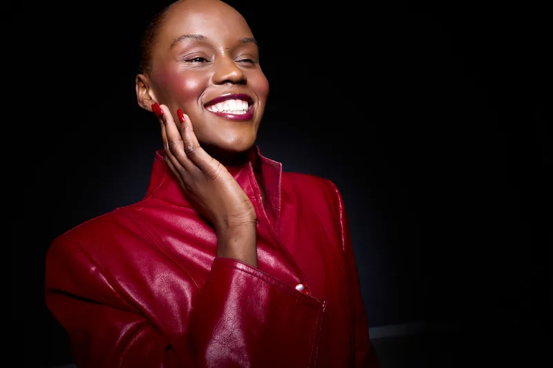 Smiling black woman with dark skin wearing a red leather jacket touches her face. Studio portrait on black background. Concept of beauty, fashion, confidence, and elegance.