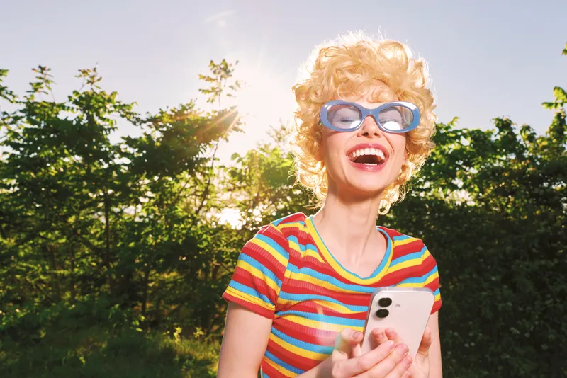 A joyful woman in a striped colorful shirt and sunglasses outdoors, holding a phone, sunshine and trees behind. A lifestyle portrait capturing carefree summer vibes and retro fashion.