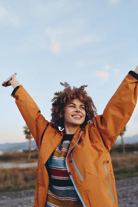 Woman with curly hair smiling outdoors, raising arms in an orange jacket and striped sweater, autumn light over countryside landscape, lifestyle moment, cheerful relaxed pose