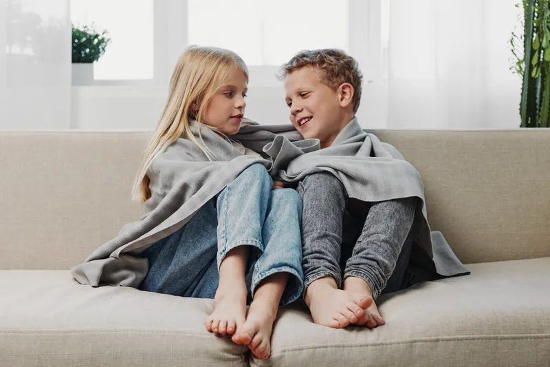 Two barefoot children wrapped in a grey blanket sit closely on a beige sofa by a bright window. The cozy indoor scene captures warmth, comfort, and sibling bonding on a relaxed day.
