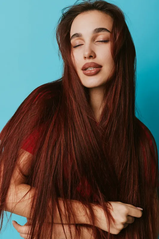 Woman with long red hair and closed eyes posing against blue background wearing red shirt with natural makeup and relaxed expression in studio portrait photography.