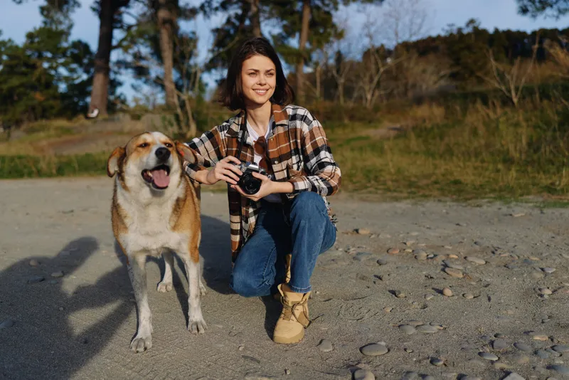 Woman crouching beside a dog on a gravel path, holding a camera ready for action, in a sunny outdoor setting with trees in the background, capturing casual photography moments for a lifestyle hobby.