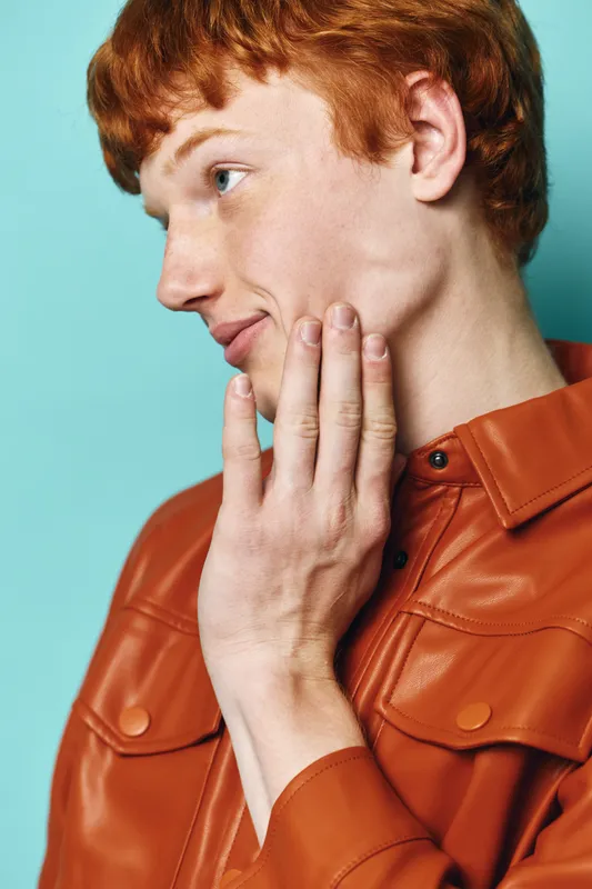 Young redhead man with clear skin touching face thoughtfully. Portrait profile with relaxed expression wearing brown leather jacket against plain background.