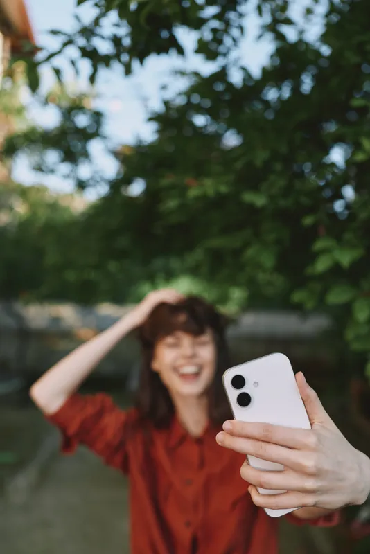 A cheerful woman in a red top takes a selfie outdoors, her smile bright as sunlight filters through leafy trees. A casual lifestyle moment captured for social media and everyday joy