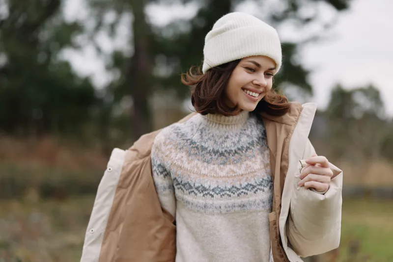 Woman in a warm knit sweater and beanie outdoors, smiling as she adjusts a light coat, in a field with soft autumn light and blurred trees.