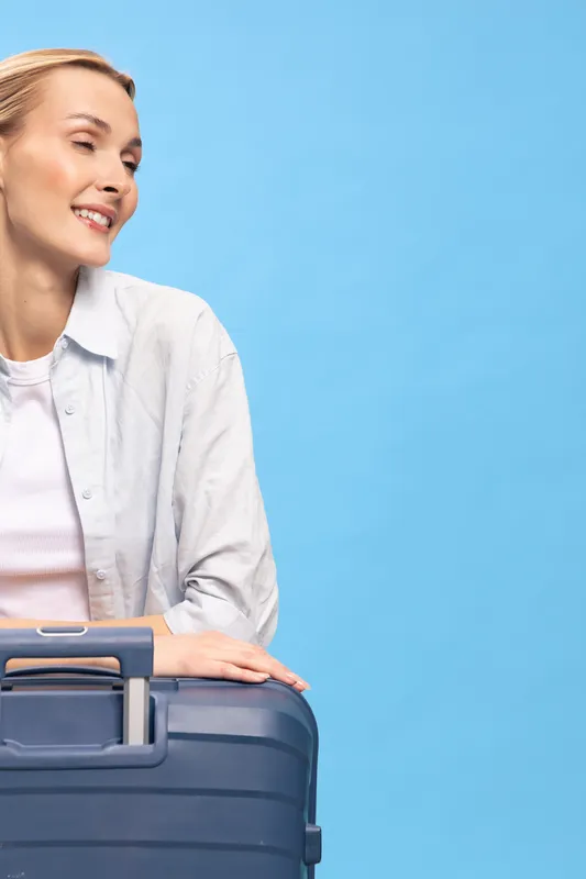 Smiling woman with suitcase stands against blue background looking sideways. Concept for travel, vacation, tourism, and summer journey with positive emotions and casual outfit.