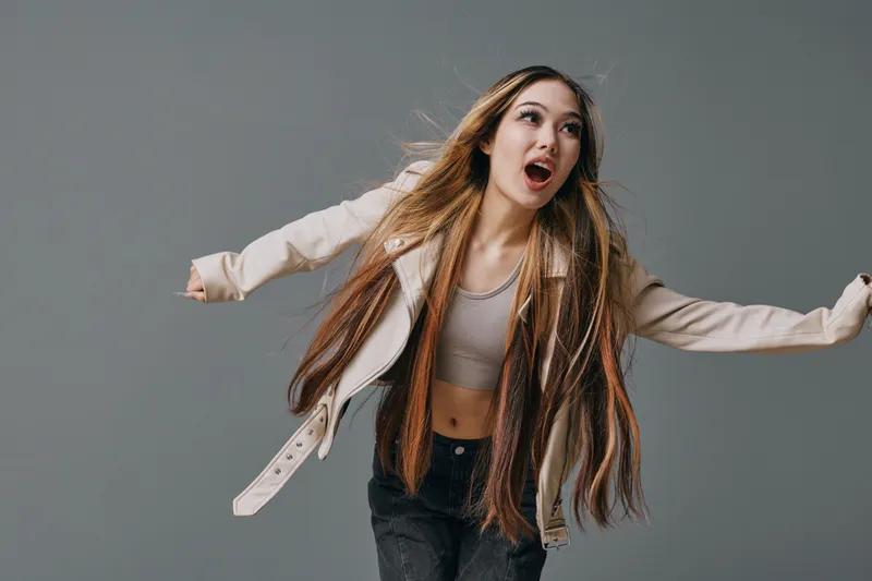 Young woman with long braided hair wearing casual clothes posing with arms outstretched looking excited on gray background in studio space.