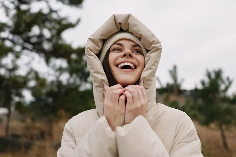 Woman in a beige hooded winter jacket outdoors, smiling warmly as she pulls the hood closer, surrounded by a muted landscape