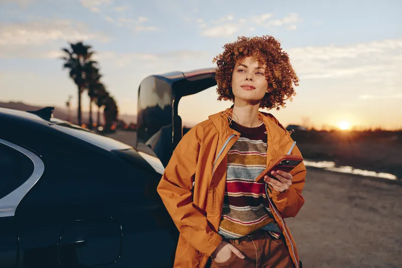 Woman with curly hair standing by a car on a road at sunset, outdoor lifestyle portrait, casual jacket and striped sweater, warm golden hour light, relaxed travel mood, open air