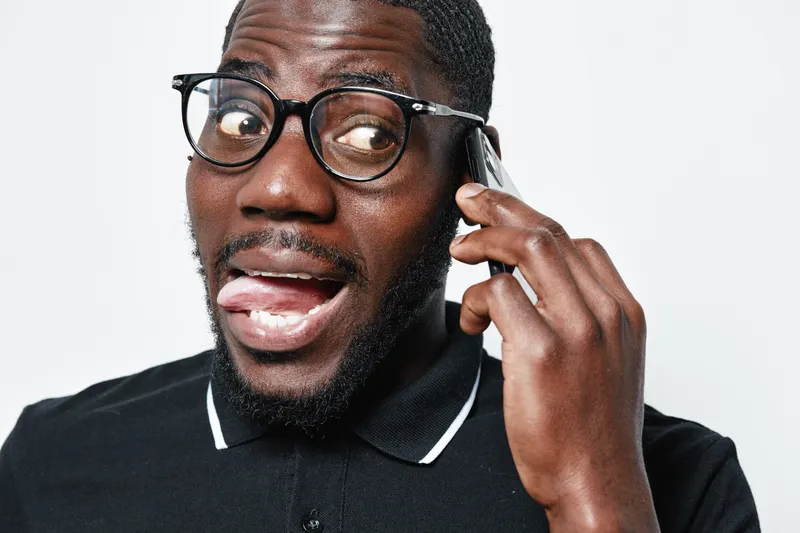 African American man with dark skin and glasses talking on smartphone with expressive face in casual black polo shirt isolated on white background for communication concept.