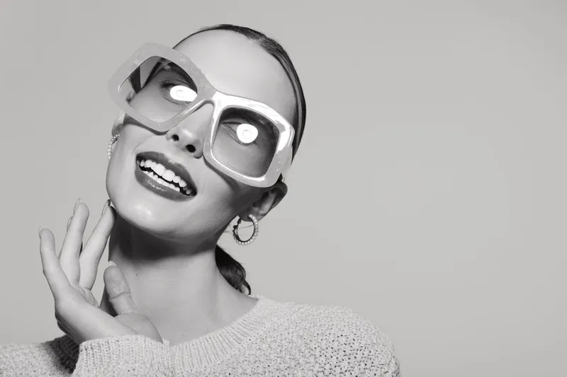 Smiling white woman with light skin and slicked back hair wearing large sunglasses and knitted sweater on grey background.