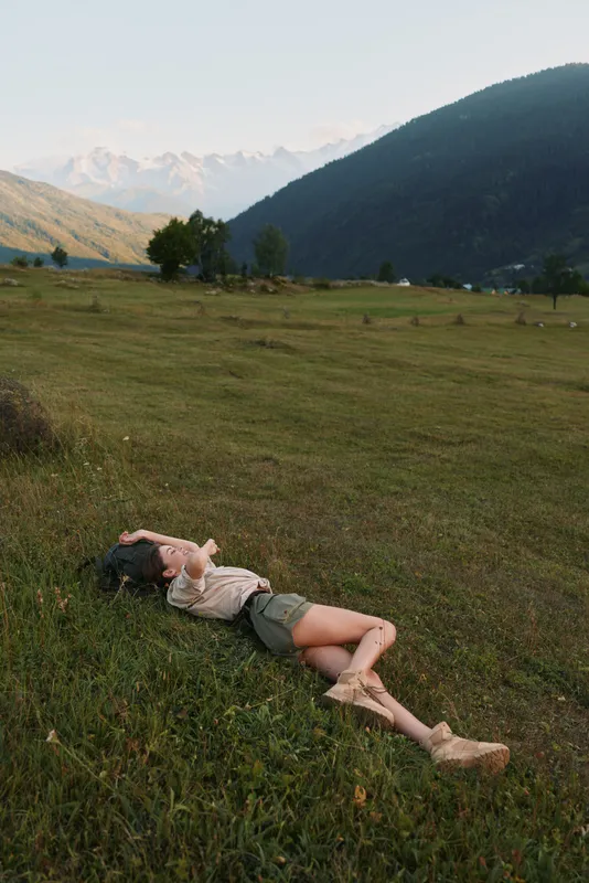 Young woman lies on green grass in open meadow surrounded by mountains under clear sky, enjoying peaceful nature and relaxed lifestyle outdoors.