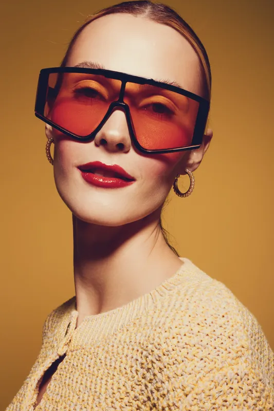 Stylish woman with oversized red sunglasses and gold hoop earrings poses against yellow background. Fashion portrait with bold makeup and trendy accessories in studio lighting.