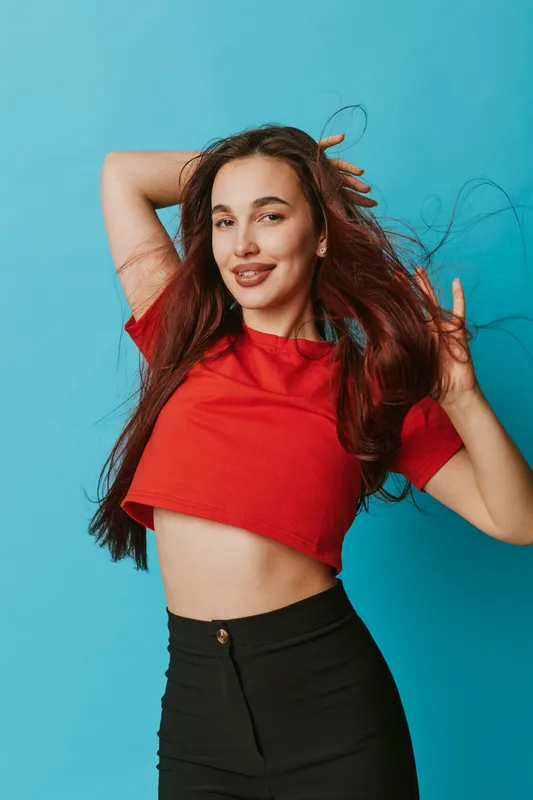 Young woman with long hair wearing red crop top and black pants posing with hands in hair against blue background. Happy female model smiling with casual style and confident expression.