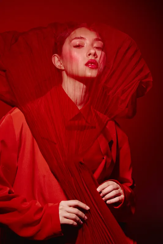 Young woman with intense expression in red dramatic pleated fabric posing with vibrant makeup stunt in creative beauty fashion editorial studio shot with moody lighting concept.