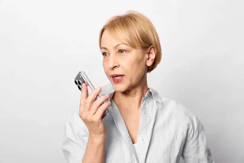 Senior elderly blonde woman holding smartphone, concern on her face as she looks aside, serious communication about technology, indoor portrait on plain light background