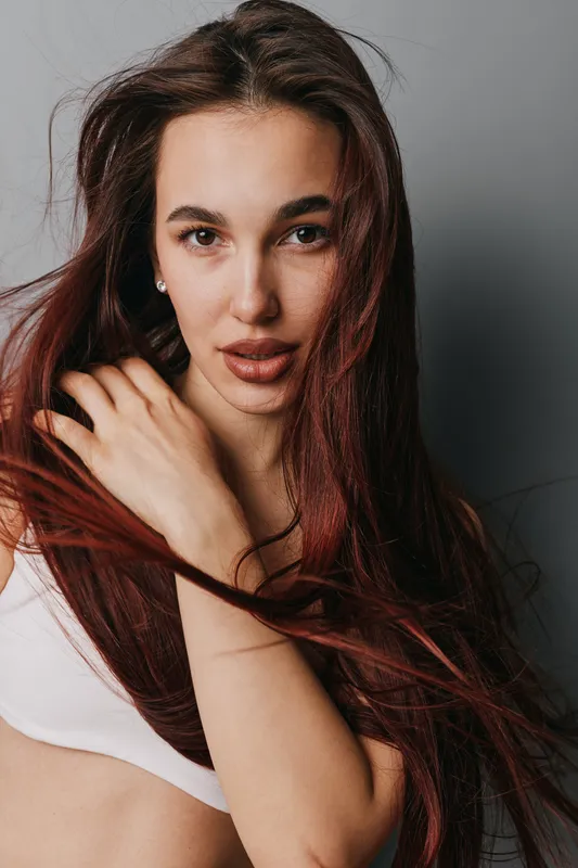 Young woman with long red hair posing in white top against gray background. Closeup portrait with natural makeup and confident expression, beauty and fashion concept.