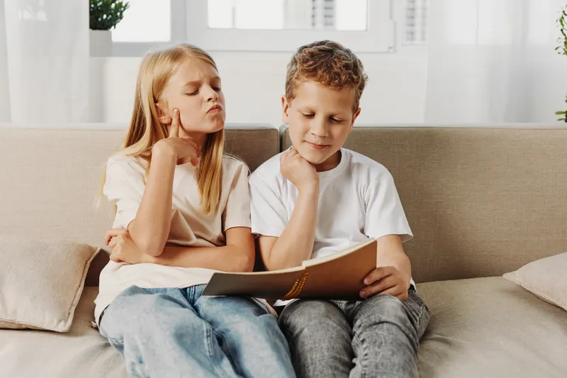 Two children sitting on a beige couch reading a book together in a bright living room. The girl looks thoughtful while the boy is focused on the pages. Concept of learning and childhood.