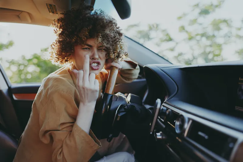 A young woman with curly hair sits in a retro tinted car, sticking out her middle finger in a bold gesture, capturing a candid moment of rebellious attitude.