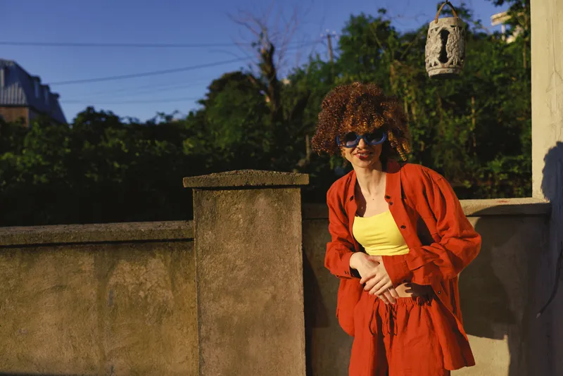 A stylish woman with curly hair in a bright orange outfit stands beside a concrete wall outdoors, sunlit and relaxed. The image conveys lifestyle, fashion, and confidence through a casual pose