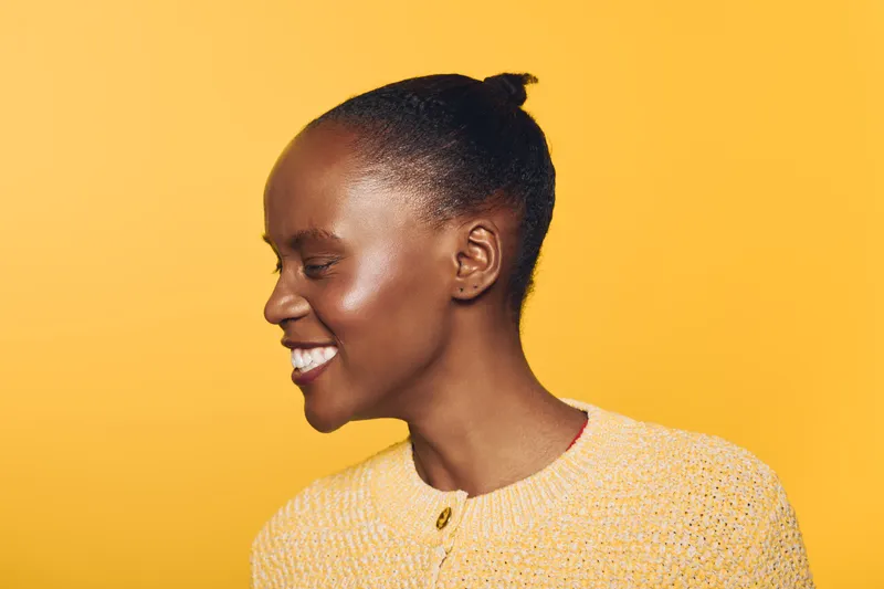 Smiling African woman with dark skin and natural hair tied back wears yellow sweater, posing sideways on bright yellow background. Concept for happiness, beauty, and natural style.