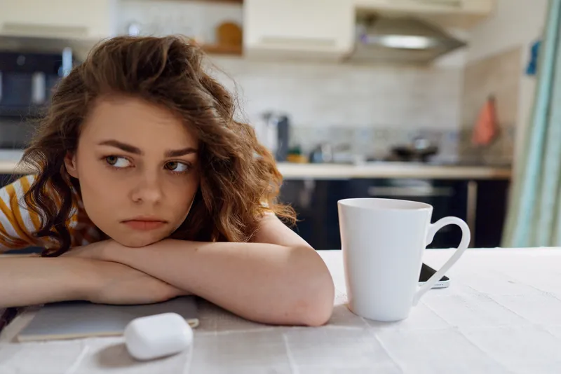 Contemplative woman at home, resting her head on a kitchen table, soft light, mug nearby, casual lifestyle moment, cozy domestic atmosphere, everyday relaxation